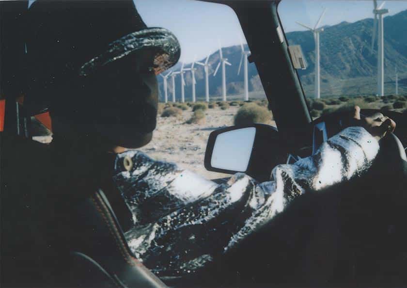 backlit person sitting in driver's seat of a vehicle, with a line of mountains and windmills visible through the windows