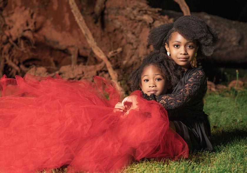 young black girls wearing red and black, sitting on the grass with upturned tree roots behind them