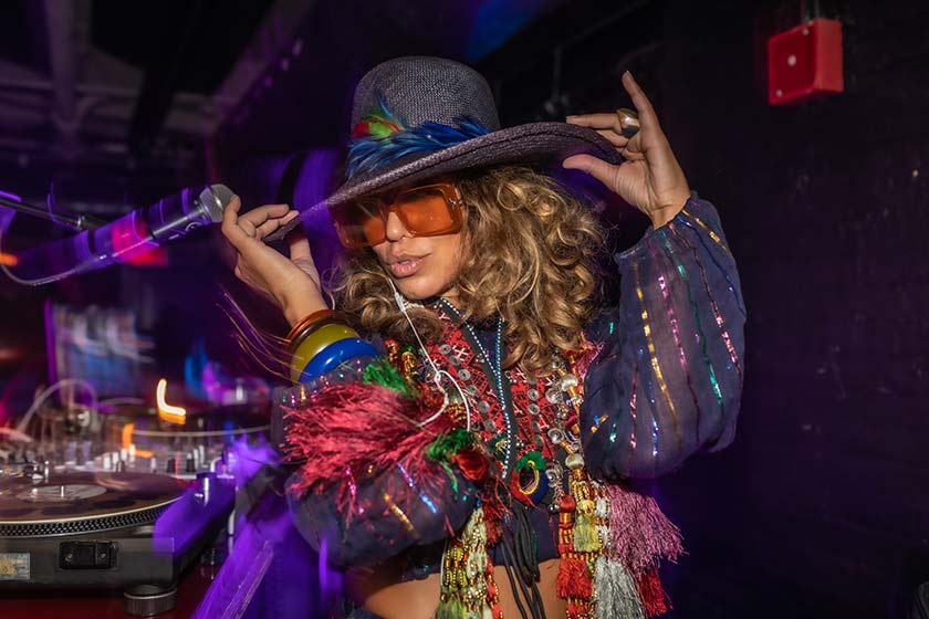 black woman posed holding her hat brim, wearing large sunglasses and colorful fringed clothing