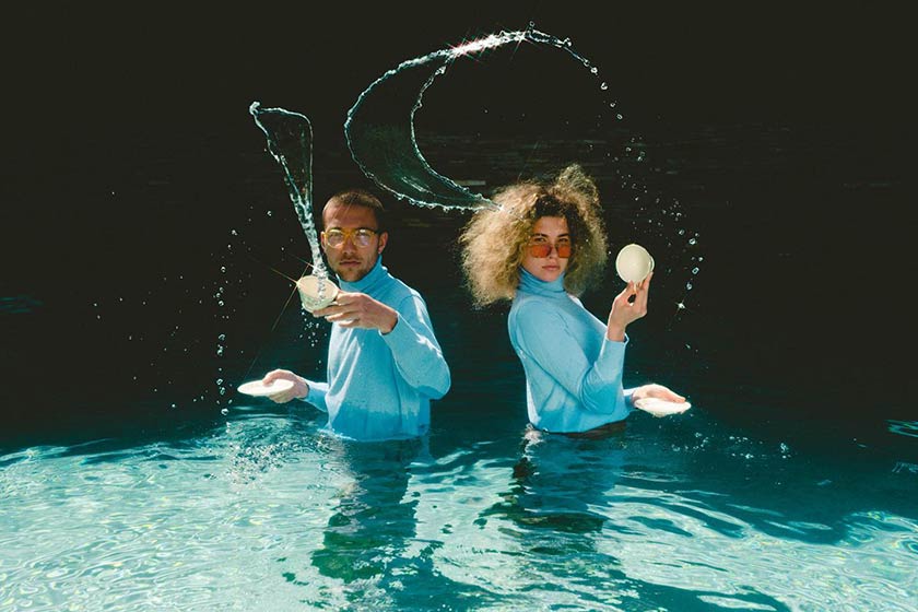 two people standing up to their waists in water, wearing blue shirts and flinging glittering strips of water with teacups