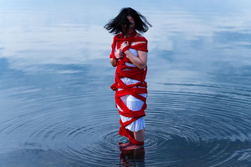 woman standing in water wearing a white dress, her entire body bound by a long red strip of fabric