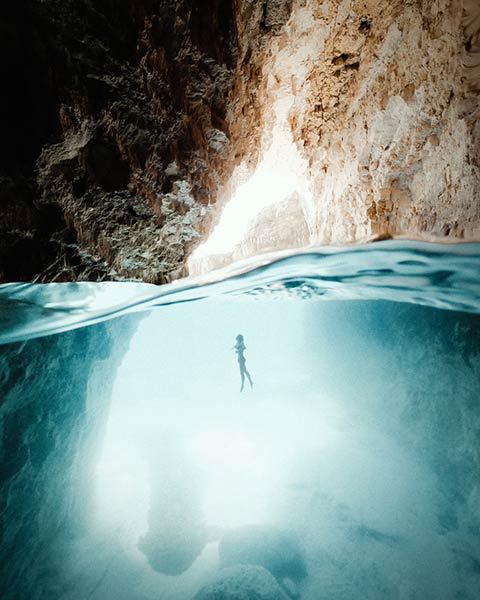 a backlit figure swimming up toward the surface of the water, near the mouth of a cave