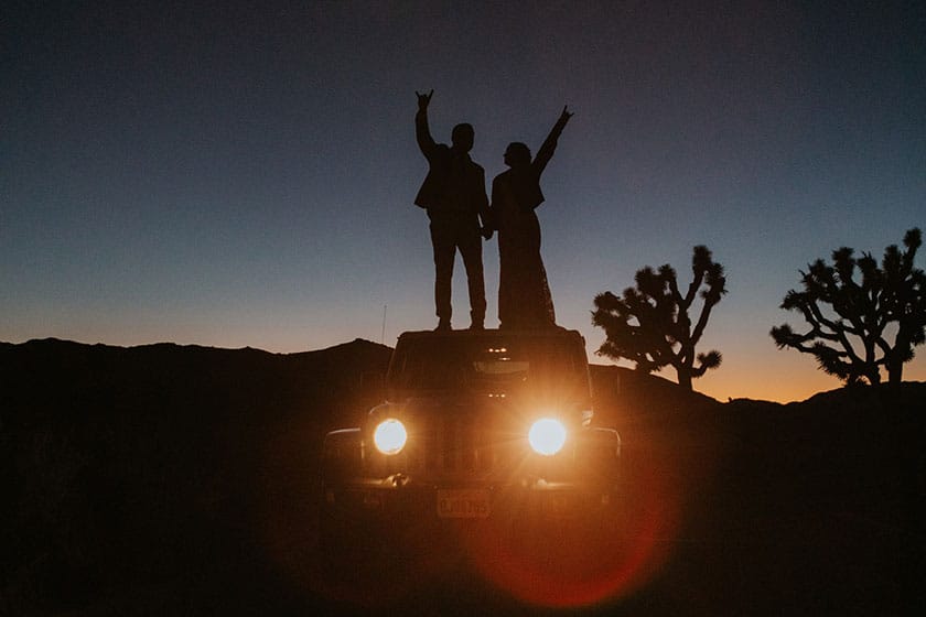 wedding couple standing silhouetted atop a jeep with glowing headlights at dusk