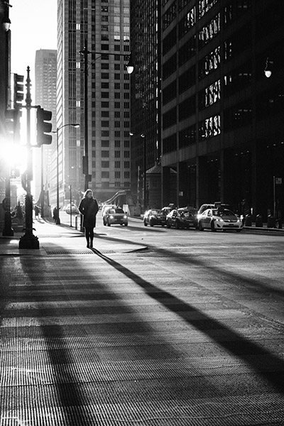 black and white street photography with a sun flare and long shadows cast by a pedestrian in the crosswalk and signposts between the skyscrapers
