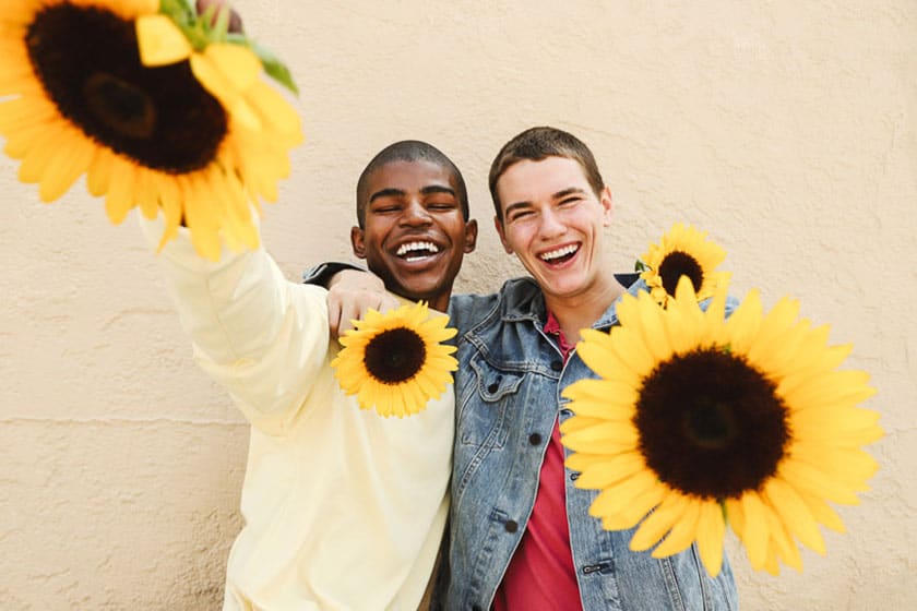 two people with their arms around each others shoulders, smiling and holding sunflowers