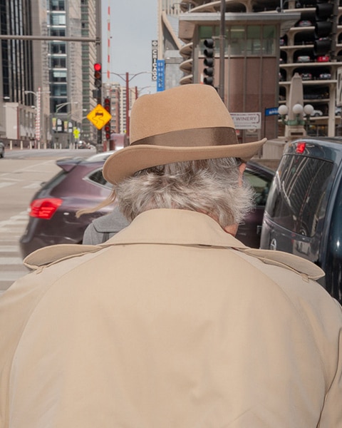 image of person wearing a felt hat and tan trench coat from behind, a city intersection in the background