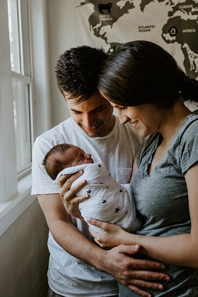 mother and father holding swaddled newborn near a window