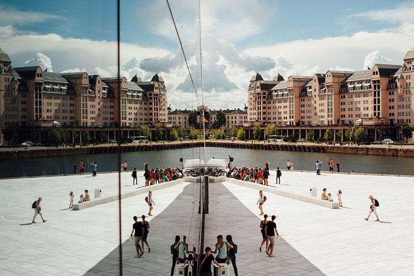 image of people walking in an outdoor plaza by a river in a city, and its reflection on a smooth building nearby.
