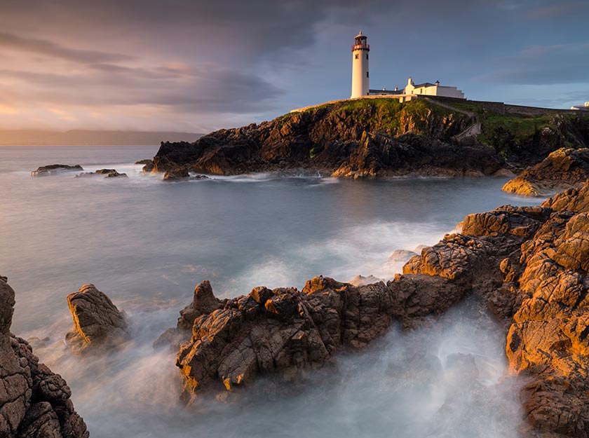 long exposure of a rocky ocean coastline and lighthouse at sunset
