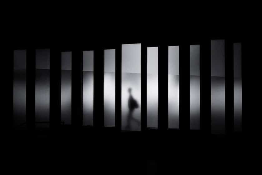 black and white image showing tall, narrow rectangles of light glowing in a dark space, with a shadow of a person walking past the central one.