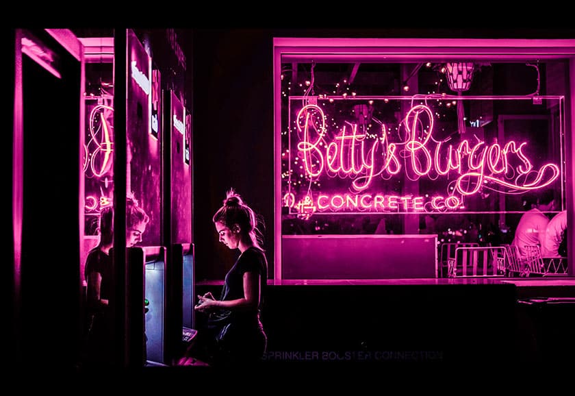 image of dark pink neon sign for Betty's Burgers, reflecting on a woman standing by an ATM nearby