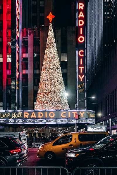 night street photo of radio city music hall with the neon lights and a seasonal tree made of tiny white lights