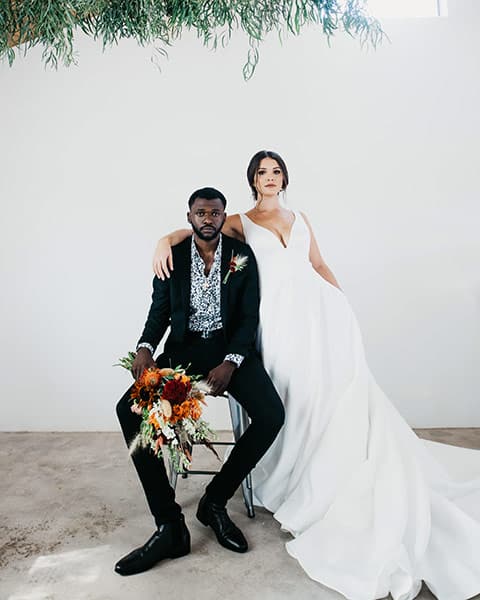 wedding portrait of groom sitting while holding the bouquet with bride leaning against him, her arm draped across his shoulders