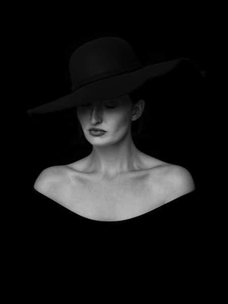 black and white portrait of woman wearing black dress and hat, her shoulders and face standing out in contrast to the dark background