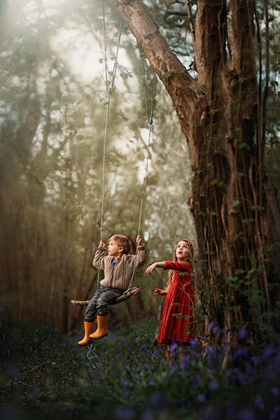 Image of young girl pushing a young boy on a swing hanging from a tree in the woods, with bluebells on the ground below them