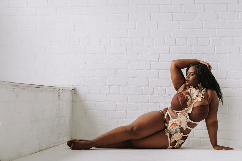 curvy black woman in lingerie posed languorously on the floor in front of a white brick wall, one arm behind her for support and the other raised, her hand resting on her hair.