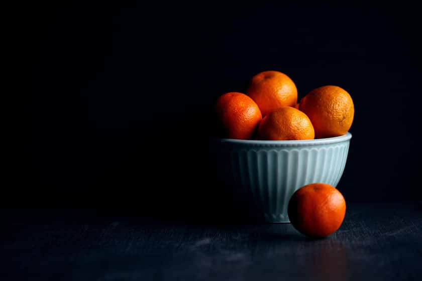 moody still life image of a pale blue ribbed ceramic bowl brimming with oranges, one orange on the surface beside the bowl, in front of a dark shadowed background