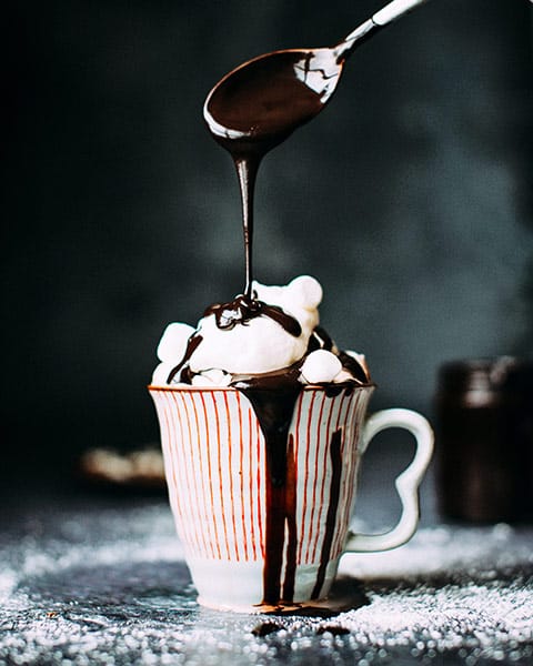 red and white striped mug mounded with whipped cream and marshmallows, and a spoon above it drizzling rivers of melted chocolate on top and down the side