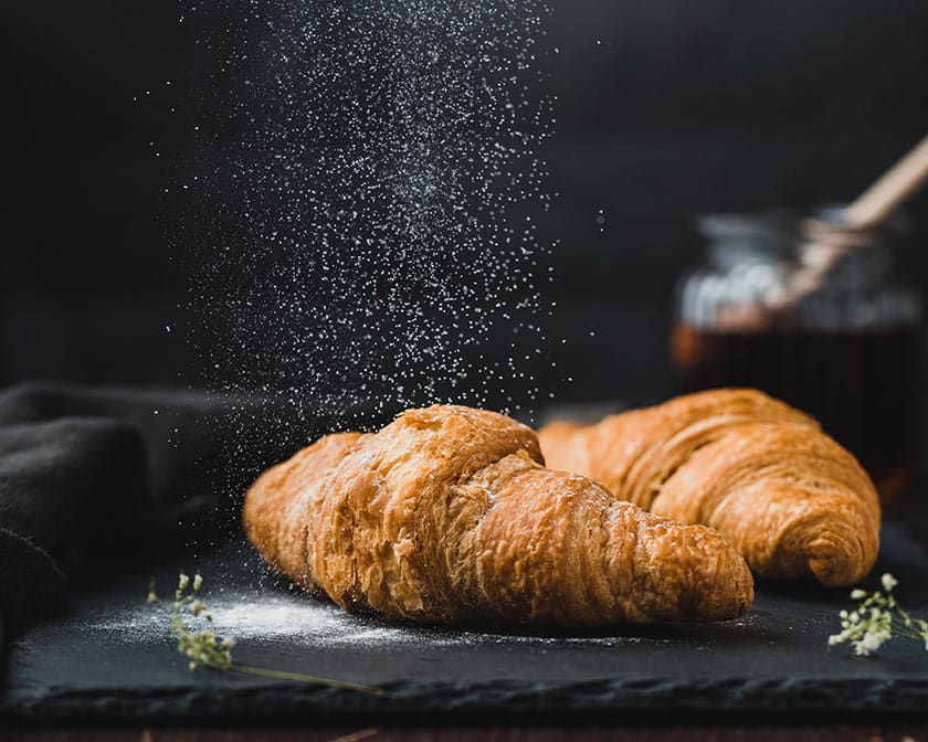 still life food photograph of two croissants on a slate cutting board, powdered sugar drifting down onto them from the top of the image