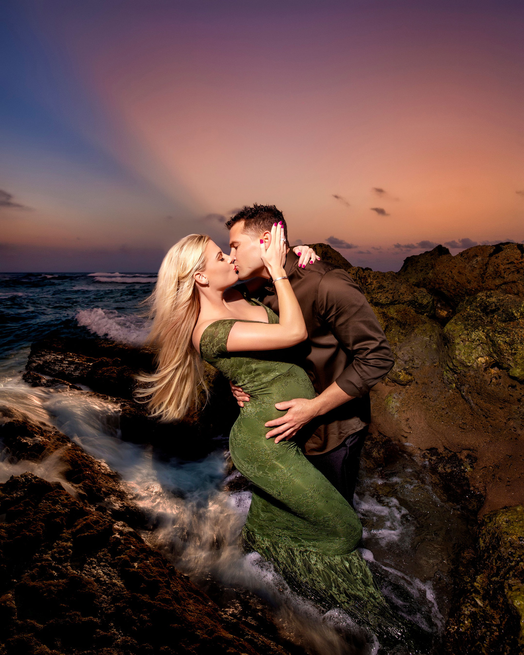 A pregnant woman in a green dress and a man in a brown shirt kiss on a rocky beach with waves crashing around them, set against a colorful sunset sky. The man's hand rests on the woman's baby bump. Photo by Adam Opris