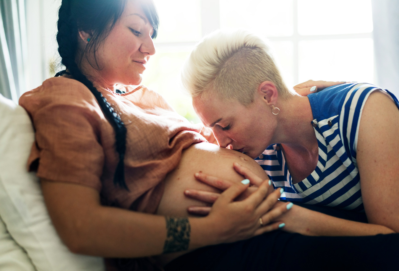 A pregnant woman with long dark hair rests against a pillow while her partner, with short blonde hair, kisses her pregnant belly. The pregnant woman is wearing a peach-colored shirt and her partner is wearing a blue and white striped shirt.