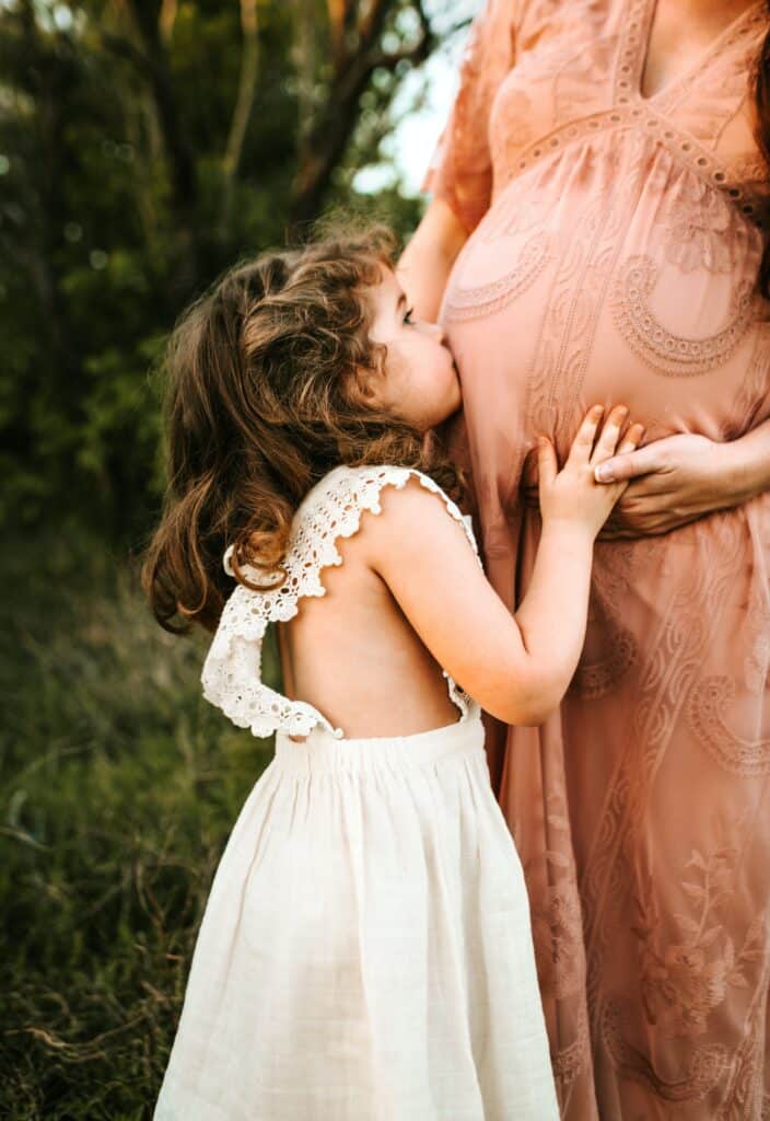 child in ivory lace pinafore dress kissing the pregnant belly of her mother