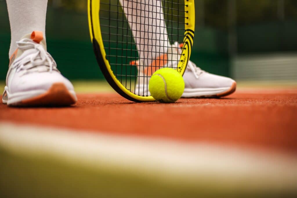 feet in white sneakers with a tennis ball resting beside the top of a tennis racket