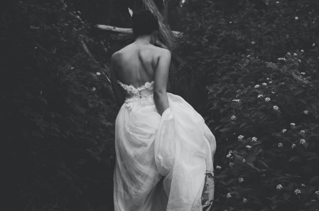moody black and white image of a bride in a strapless dress walking between flowering plants, holding her train