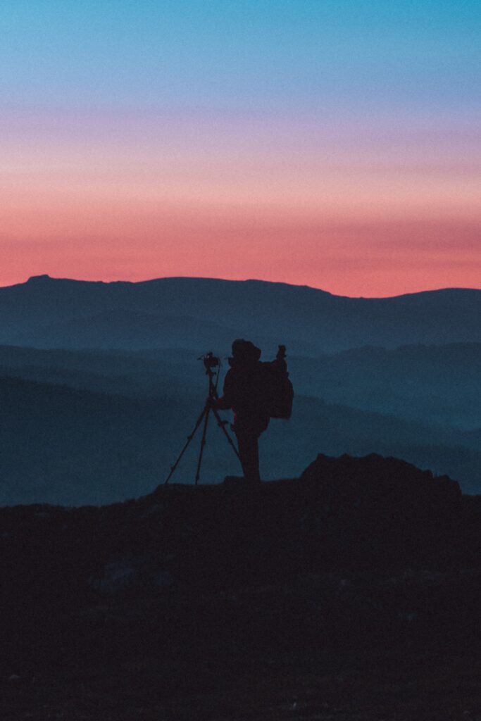 a person carrying a backpack standing with a camera on a tripod on top of a mountain at sunset