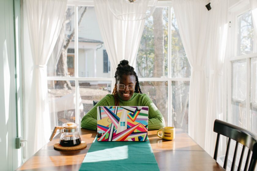 black female sitting at wooden table with a coffee pot, mug, and using a colorful laptop