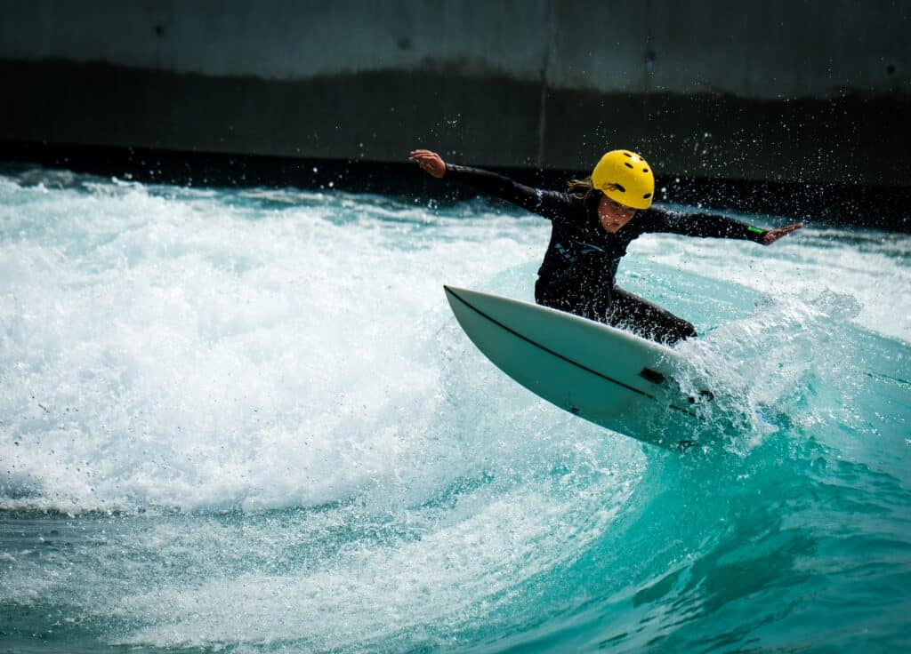 kid surfer wearing black wetsuit and yellow helmet riding a wave on a white board in a wave pool.