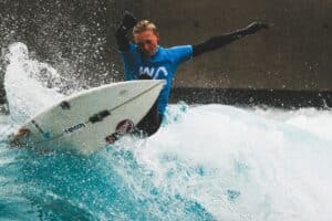 Surfer in blue wetsuit riding a wave on a white board at The Wave, a wave pool in Bristol. The Wave is an artificial, inland wave pool where you can learn to surf.