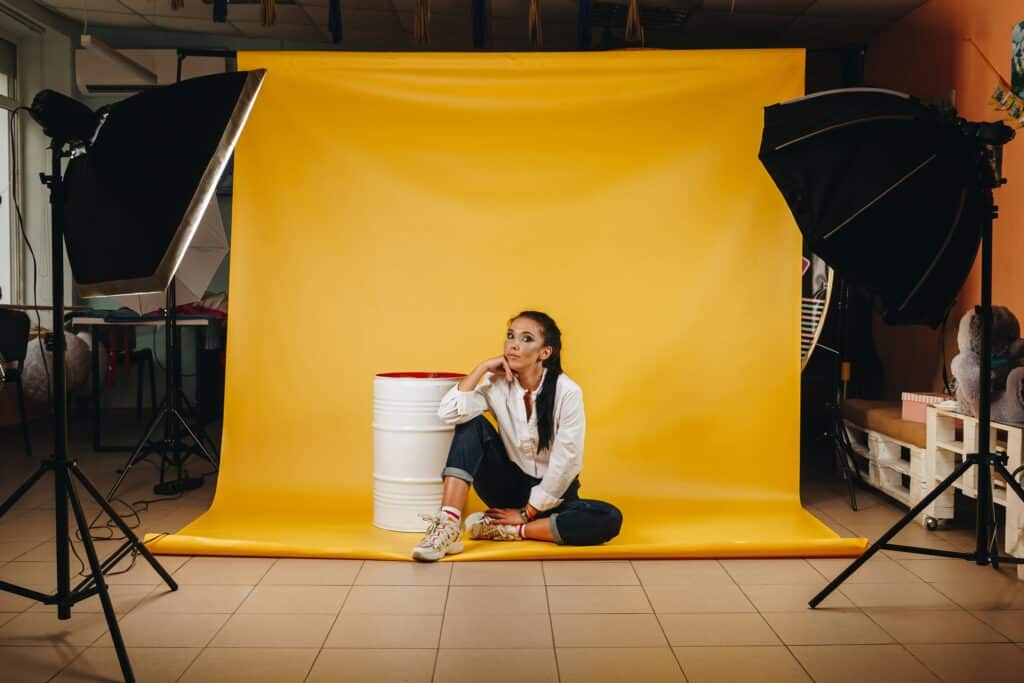 woman sitting on a yellow backdrop in a photography studio with a studio light and softbox or umbrella on either side of her, at a 45 degree angle