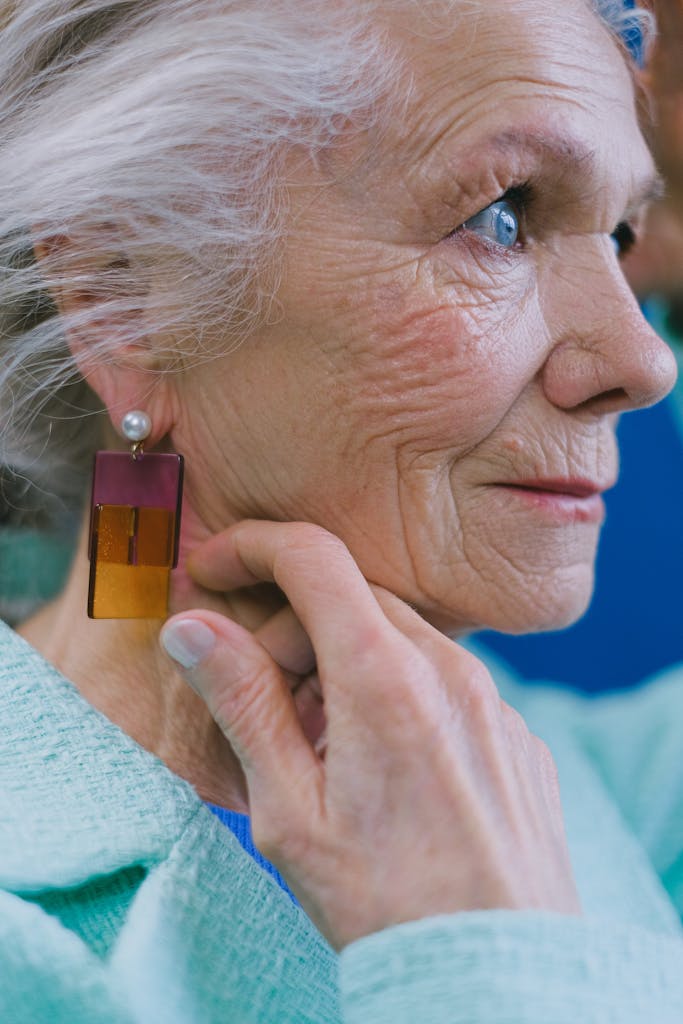 Older woman with blue eyes, silver hair, and softly wrinkled skin touching her jawline near a dangling earring and looking away from the camera.