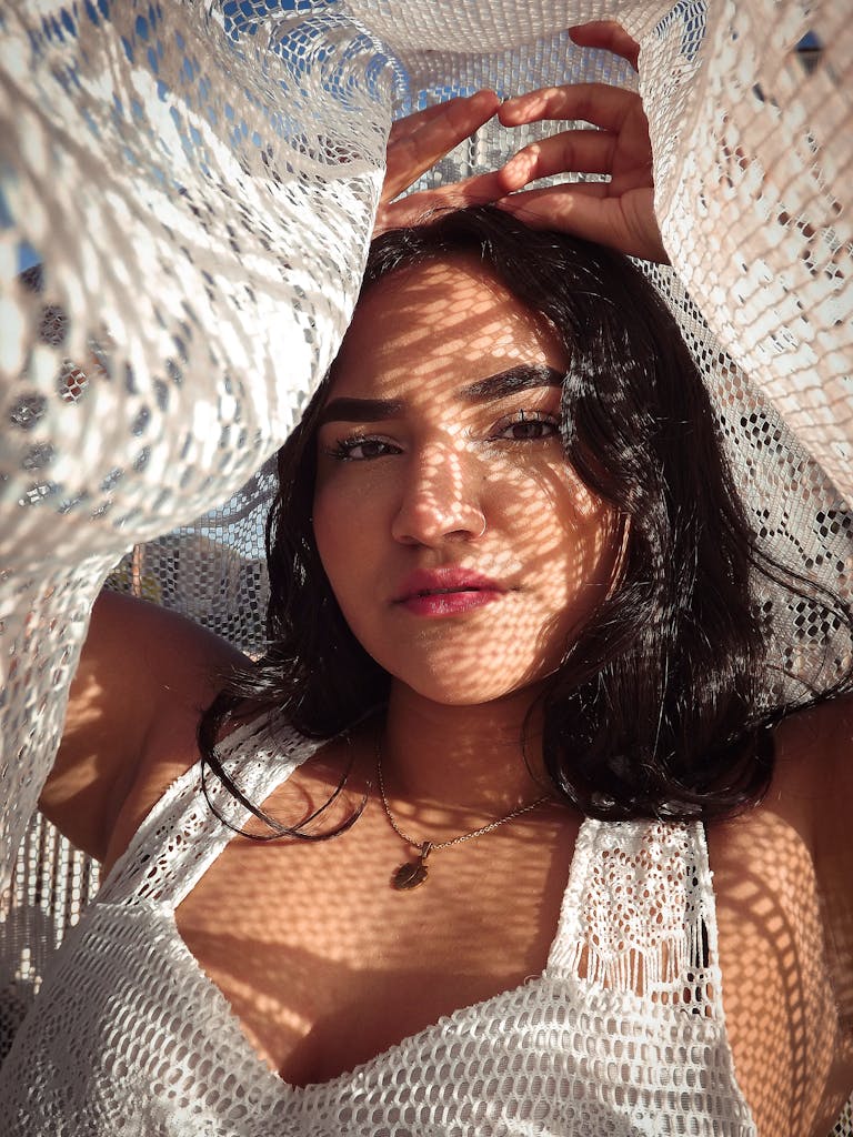 Woman in White Knit tank top with a white lace cloth over her head and the camera lens, the sunlight filtering through it and making patterns of shadow and light on her skin.