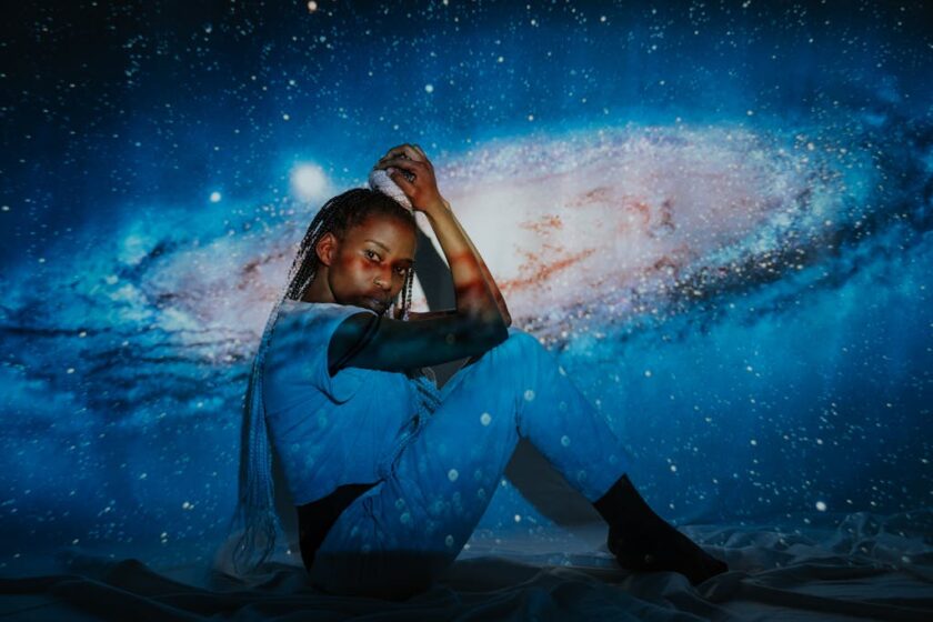 Woman Sitting with knees bent, elbows resting on her knees, and hands gently grasping the braided bun of hair on her head while she looks at the camera. A scene of a galaxy is projected onto her and provides the light for the image.