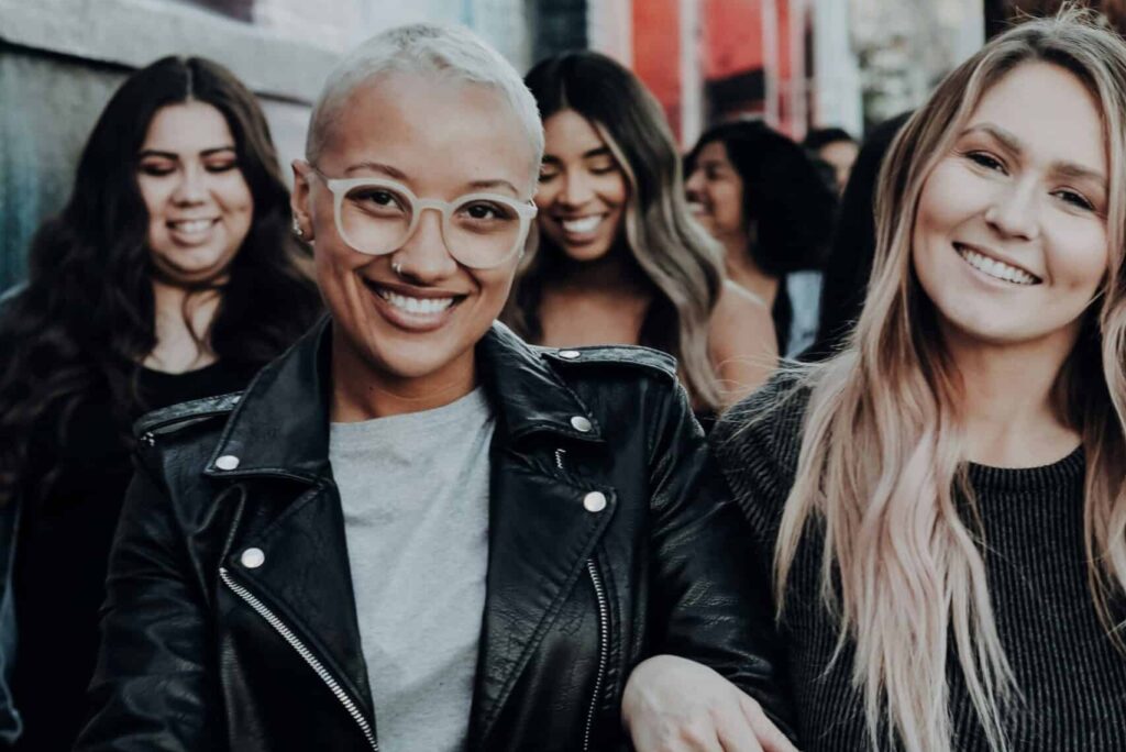 Black woman with short hair wearing glasses and smiling wearing a black leather jacket walking with arms linked with a white woman with long hair and black shirt. Several more BIPOC women are visible walking behind them in a group.
