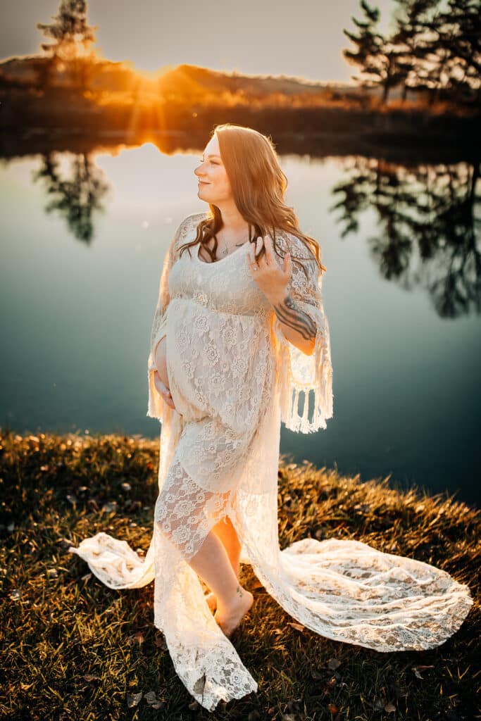 Maternity portrait of woman in lace dress standing beside a lake at sunset by Amy Helt Photography