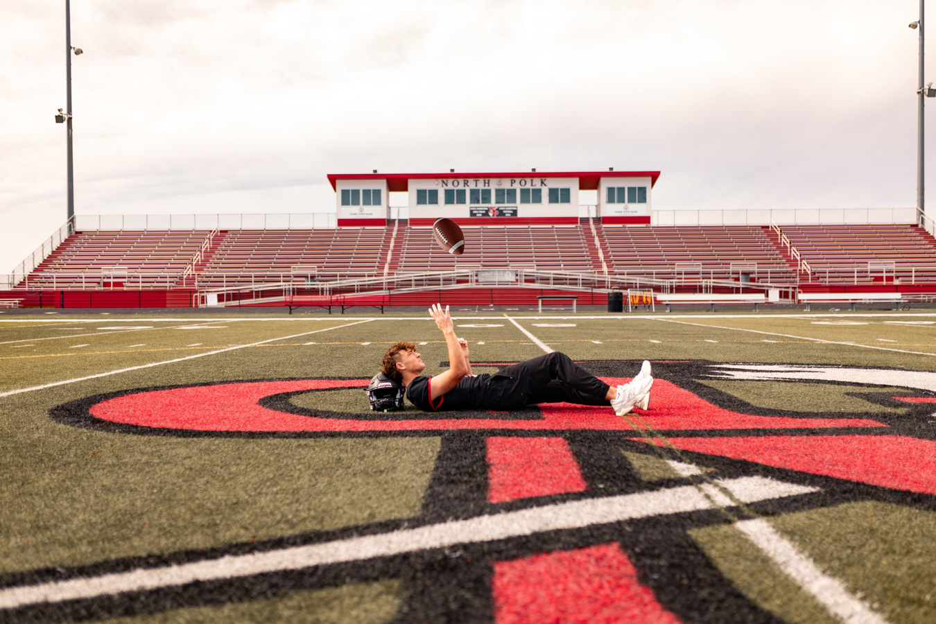 A young football player lies on his back on the red and black logo of a turf field, tossing a football in the air with empty red bleachers and a press box in the background.