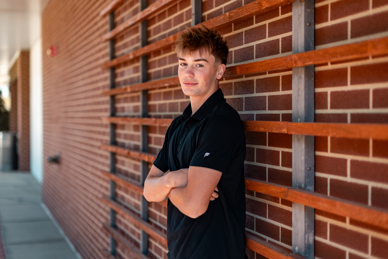 A young man with messy brown hair stands with his arms crossed, smiling slightly while leaning against a brick wall with a wooden trellis structure.