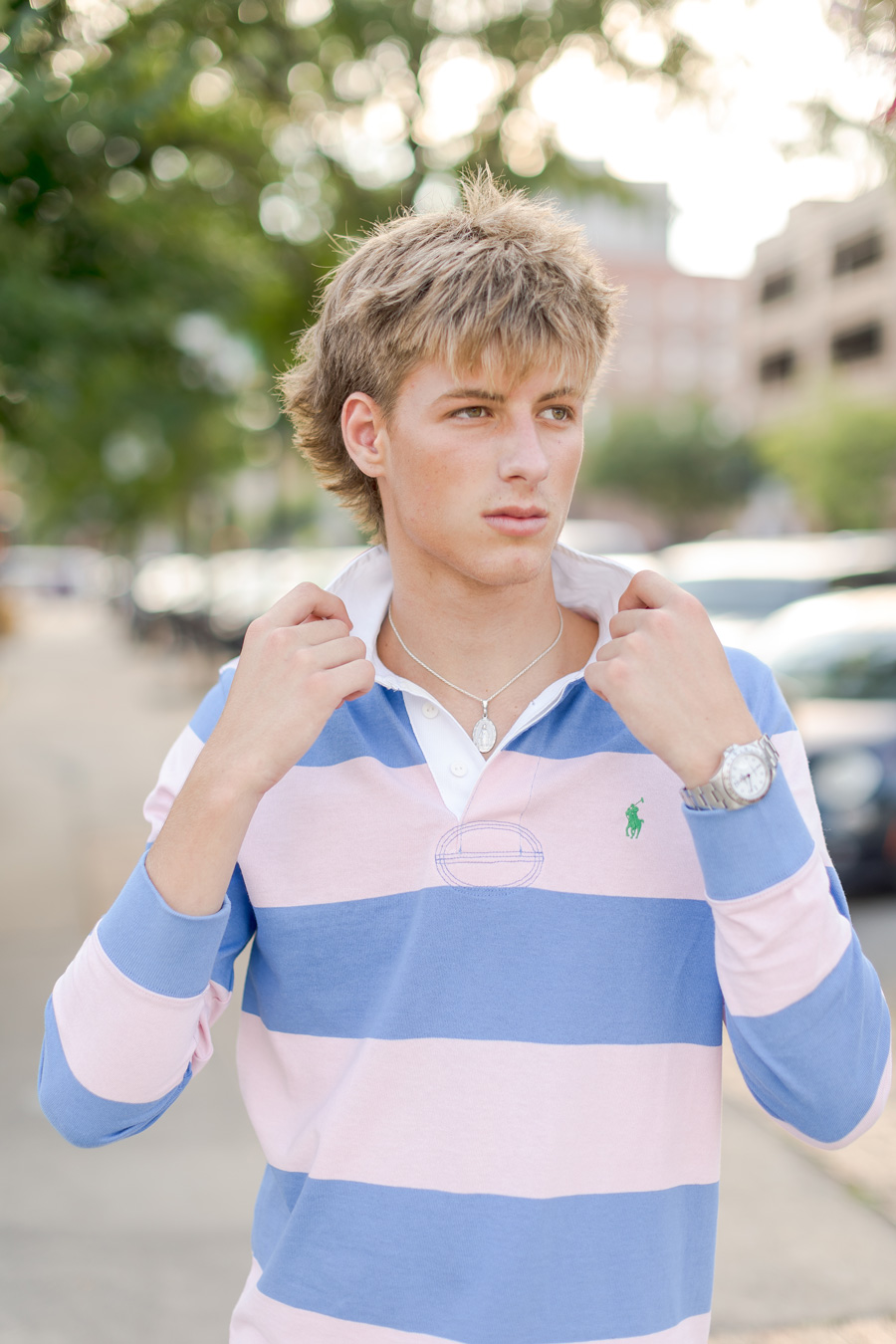 A young man with a blonde messy haircut stands outdoors, adjusting the collar of his pink and blue striped long-sleeve polo shirt while wearing a silver necklace and watch.