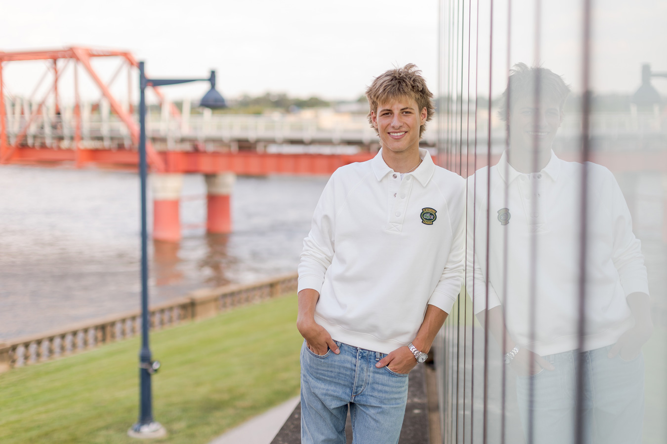 A smiling young man in a white long-sleeved polo shirt and blue jeans leans against a reflective glass wall, with his reflection visible beside him and a red bridge over a river in the soft-focus background.
