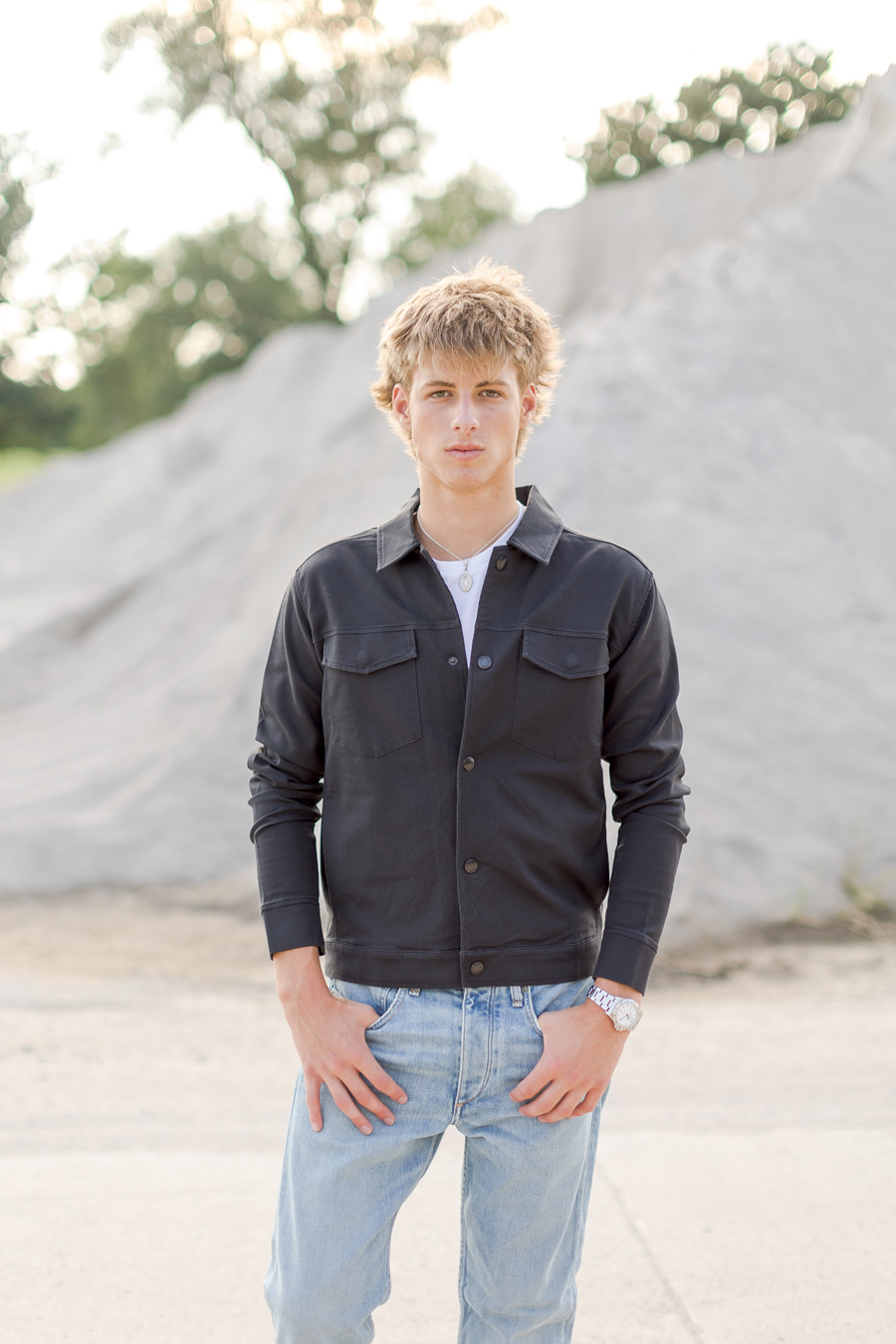A young man with messy blonde hair stands outdoors with his hands in his pockets, wearing a black button-up jacket over a white t-shirt and light-wash blue jeans, with large mounds of sand in the background.