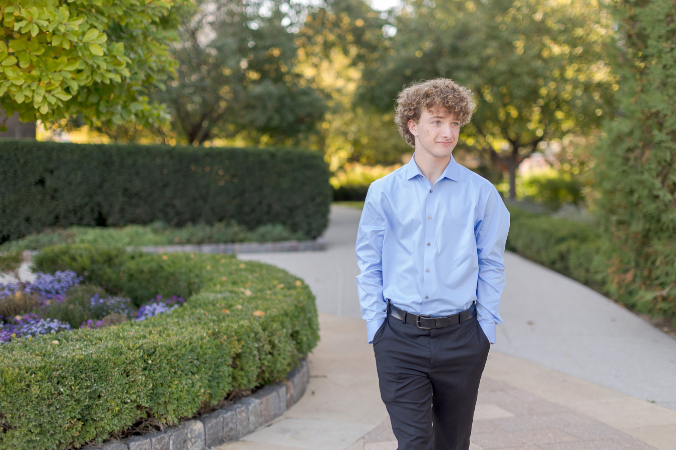 A young man with curly light brown hair walks along a paved path in a park, wearing a light blue button-down shirt and black trousers with his hands in his pockets.