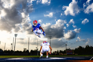 football player in white jersey crouched on ground while a teammate in a blue jersey jumps over his back