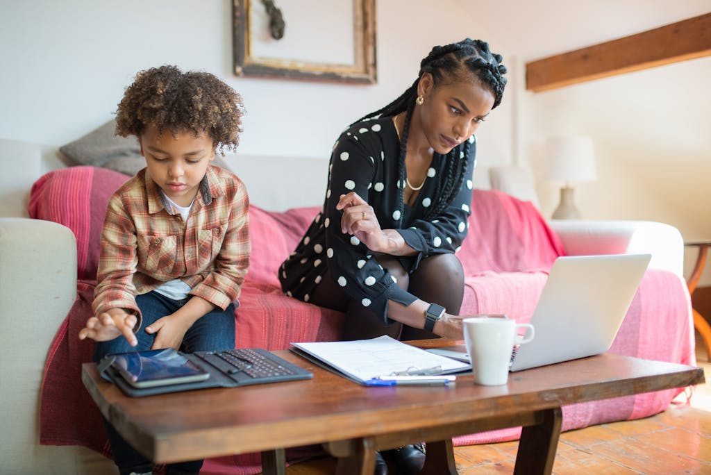 mom working on laptop at couch beside child using tablet