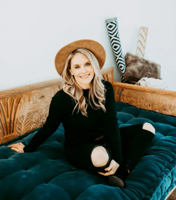 Smiling woman in brown hat and black outfit sits on teal velvet daybed