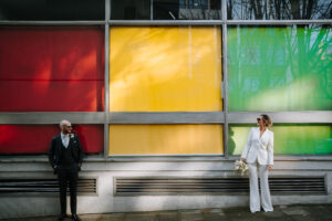 Wedding couple wearing suits in front of a colorful building. photo by Steve Bridgwood