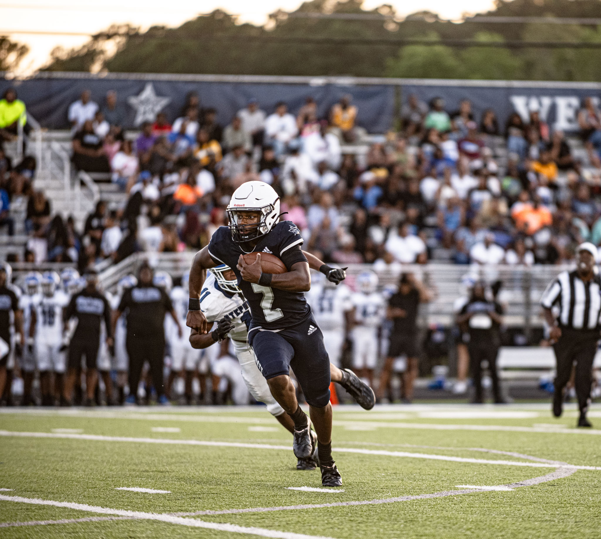 Football player running with the ball, pursued by a defender, during a game. Crowd in background
