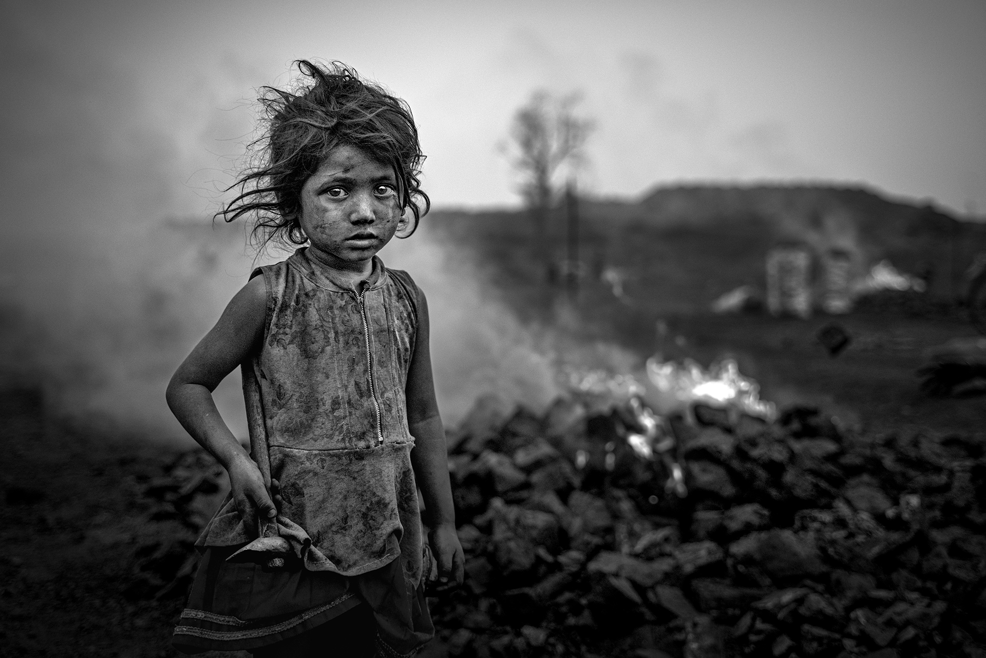 Black and white photo of a coal-dust covered child standing near a fire, looking directly at the camera. Photo by Sourav Das.
